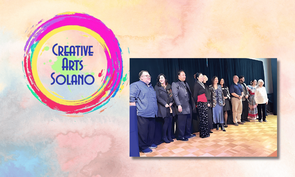 The image features the logo of Creative Arts Solano, characterized by a vibrant, multicolored paint splash forming a circle with the organization's name. Adjacent to the logo, there is a photograph of a group of individuals standing on stage during an awards ceremony. They are lined up side by side, smiling towards the audience, dressed in formal attire. This setting suggests a celebratory and formal event, likely associated with the presentation of an award or recognition.